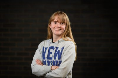 Photo of a white girl with blonde hair blue eyes wearing a grey jumper with New York in big bold dark blue writing smiling arms crossed in front of a black bricked background