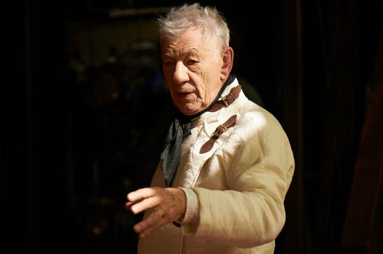 Dressed in a fencing outfit, Ian McKellen holds up his hand, gesturing to somebody off camera.