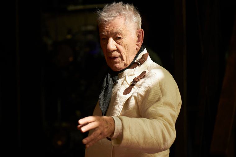 Dressed in a fencing outfit, Ian McKellen holds up his hand, gesturing to somebody off camera.