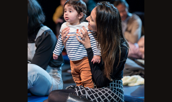 A lady is wearing a black top and a patterned skirt. A toddler in a stripy top stands on her lap.