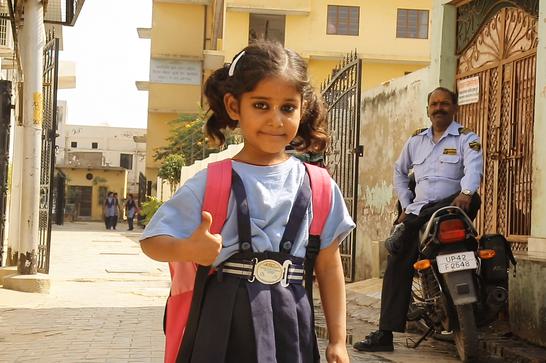A small girl wearin a red backpack gives a thunbs up to the camera. A man in police uniform rests on a motorbike in the background.