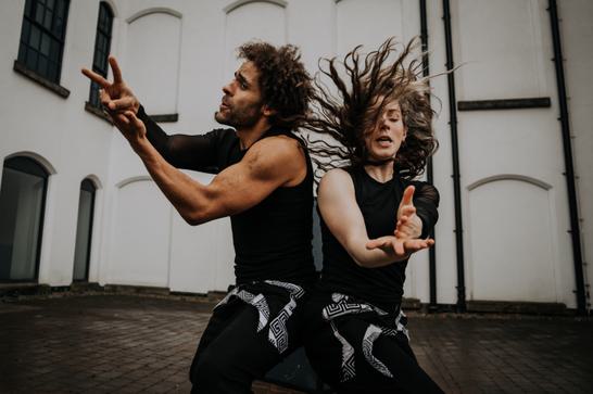 2 dancers stand back to back with their hair moving and arms outstretched.