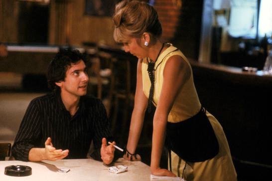 A man sits at a table holding a cigarette. A waitress dressed in yellow stands next to him listening as she rests her hands on the edge of the table.