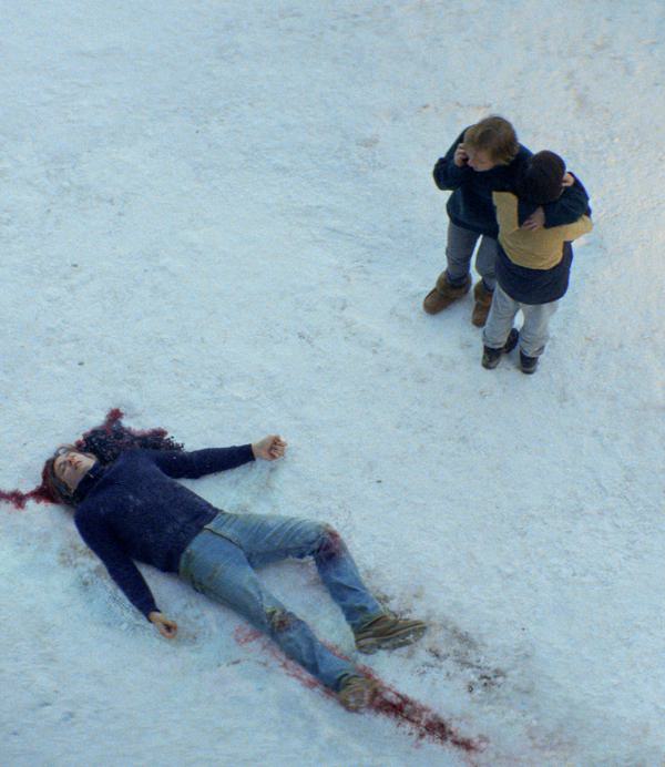 A man's body lies sprawled in the snow, blood pooled around his head. A woman and her young son stand next to it. The boy is looking away and the mother is on a mobile phone.