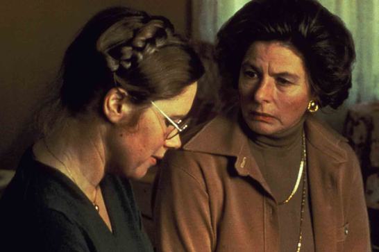 A woman in glasses with braided hair looks down as an older woman looks at here disapprovingly.