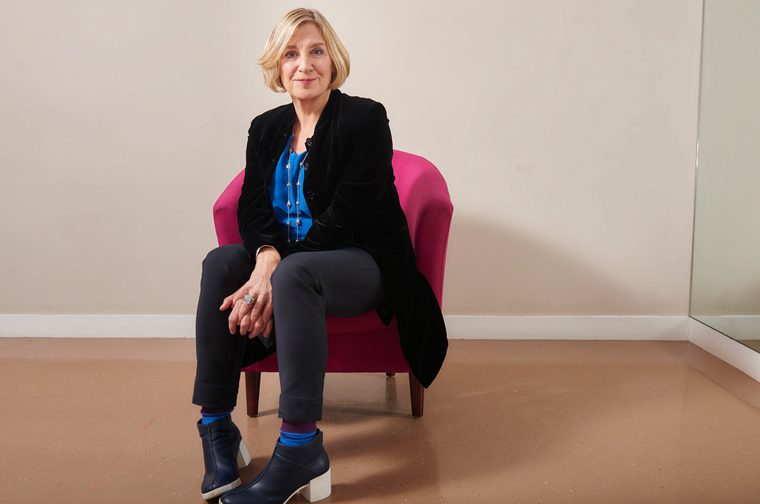 A woman in her forties (Victoria Wood) with short blonde hair wearing a black blazer with a blue blouse sits on a dark pink sofa chair. She clasps her hands between her legs as she smiles at the camera.