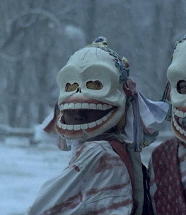 Two young children (a boy and a girl) stand in a snowy forest wearing disturbing masks, looking directly at the camera.