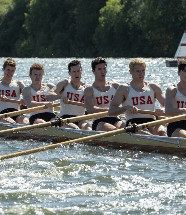A male rowing team in white vests with the letters USA imprinted on them row through a lake.