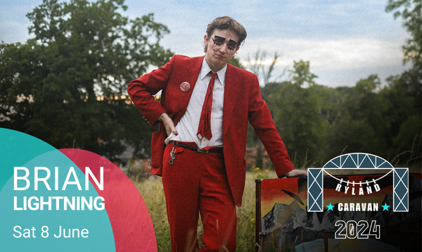 Brian Lightning stands in a red suit with heavy makeup around their eyes in a field next to their painted organ.