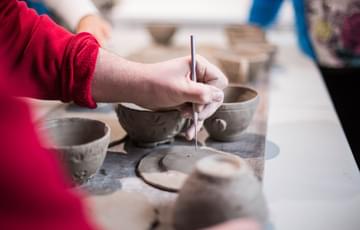 A variety of ceramic style bowls are in the centre of the image, and someone wearing a red jumper is reaching in with a tool to make surface decorations on the ceramic pieces.