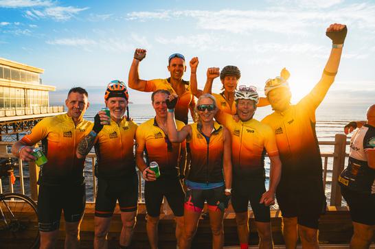 Eight cyclists wearing yellow and black outfits cheer and raise their hands, looking directly into camera. Behind them is the sea and a blue sky, with the sun shining brightly.