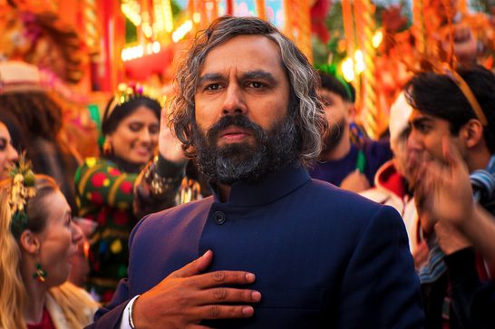 An older Asian man in with grey in his dark hair and beard looks off screen, holding his right hand on his chest. Behind him are lots of smiling people wearing festive clothing and headbands. There seems to be a carousel behind them too.
