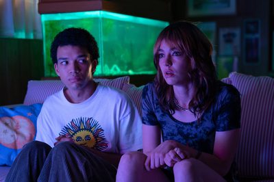 A young man looks ominously at a young woman who is staring off screen, both of them sat on a sofa. There is a neon fish tank situated behind them and they are both bathed in pink light.