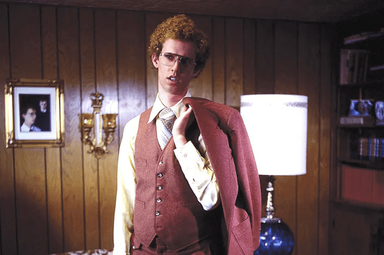 A man with curly hair and glasses (Jon Heder) is wearnig a waiscoat and holds his jacket over his shoulder in a room with wood-panelled walls and a lamp.