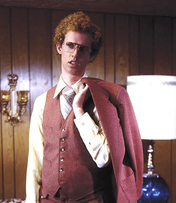 A man with curly hair and glasses (Jon Heder) is wearnig a waiscoat and holds his jacket over his shoulder in a room with wood-panelled walls and a lamp.