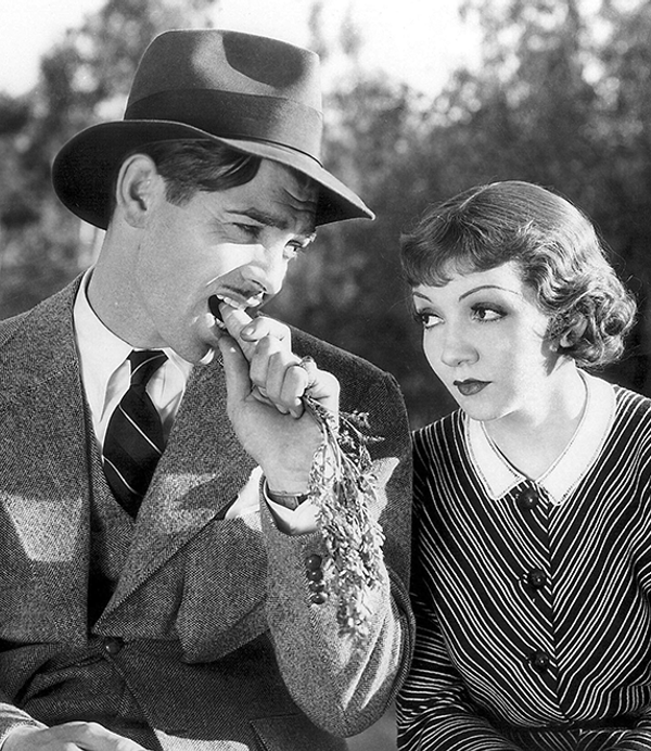 Black and white - a man in a hat bites into a carrot, sat next a woman in a collared dress.