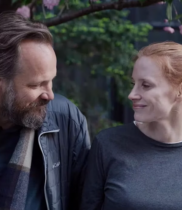 A man on the left with long hair and a beard wearing a scarf (Peter Sarsgaard) faces a woman with red hair (Jessica Chastain) on the right - they are both smiling.