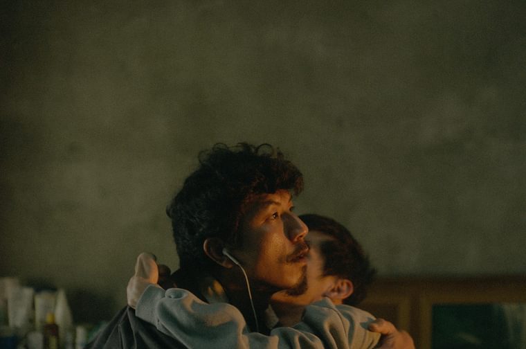 A bearded Taiwanese man wearing white ear phones hugs a younger Taiwanese man, who clutches him around his neck. The older man looks to the right of the picture, sunlight glowing on his face.