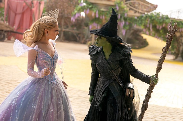 A fairy princess (Ariana Grande) wearing a large sparkly dress stands next to a witch with green skin (Cynthia Errivo) who is holding a large broomstick and wearing a large pointy witches' hat. They seem to be engaged in an argument.