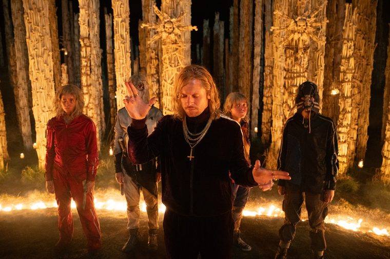 This image has a striking, ritualistic atmosphere. It shows five figures standing in what appears to be a nighttime forest ceremony. The scene is dramatically lit with flames on the ground and tall wooden posts or totems in the background, some adorned with what look like starburst decorations. The central figure in the foreground wears black clothing with chains and a cross necklace, with shoulder-length blonde hair, hands positioned as if in prayer or ritual gesture. The four figures behind are arranged in a line, with the leftmost wearing red, and the others in darker clothing. The rightmost figure appears to be holding something up.