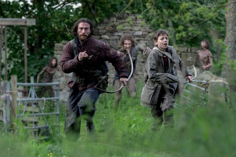 A bearded man in his forties holding a crossbow runs through a grassy field alongside a young boy, also holding a crossbow. Behind them we can see three of the Rage infected zombies racing after them.