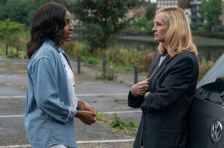 Two women are having a conversation outdoors near a car. The younger woman is wearing a denim shirt and the other older woman is dressed in a black blazer. The setting looks like a residential area with trees and buildings in the background.