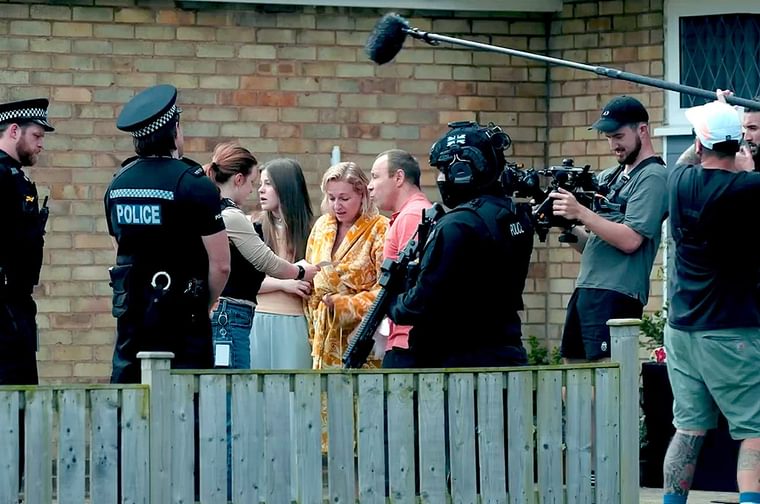 On set photo of a camera crew filming a scene with actors, two of whom are dressed as policemen. They stand in front of a brick wall with a fence to their left. We can see one camera and one boom mic.