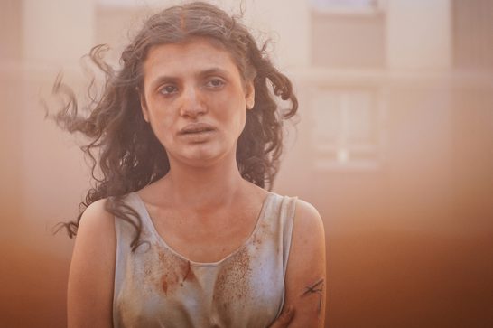 A young woman with long, curly, brunette hair stands in front of a housing tenement, surrounded by brown dust. Her face and body is caked in dust, including her white blouse, and there are sreaks of dirt on her left arm..