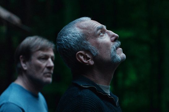 A man in his sixties (Daniel Day-Lewis) with grey hair and a trimmed grey beard and moustache stares up into the sky. A similarly aged man (Sean Bean) stands just to his left looking at him. They are standing in a wood with the wall of a small cabin to the left of the image.