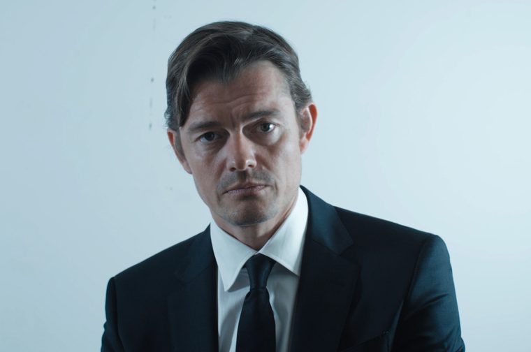 A man in his forties (Sam Riley) wearing a black suit and black tie stares morosely at the camera.