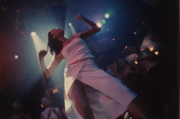 Photograph of a woman dancing in a club. She is wearing a matching white top and midi skirt, with a split in the front. There are two spotlights above her.
