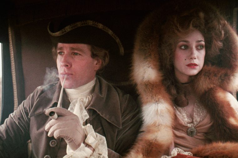 An aristocrat and a wealthy ladt sit in the back of a horse drawn carriage. The man wears a tricorn hat and smokes a long pipe, whilst the lady wears a large fur coat with the hood around the back of her head.