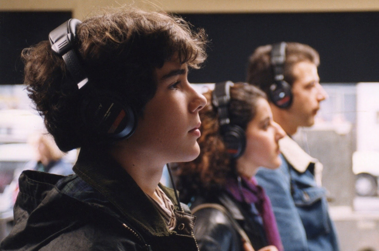 Three young people stood in a line, looking to the right. They are all wearing black headphones.