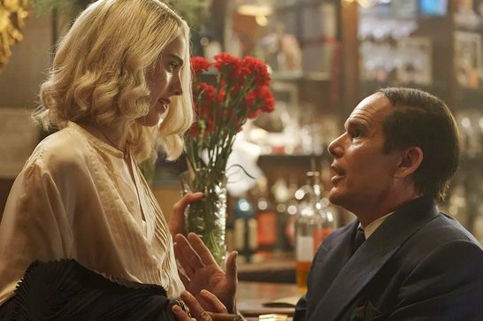 A young woman with bleached blonde hair stands next to a bar looking down at a seated man in his fifties. The man wears a blue jacket and has a slicked combover hairstyle. The woman is holding a case of red flowers and we can see various bottles of alcohols behind them stacked across the bar.