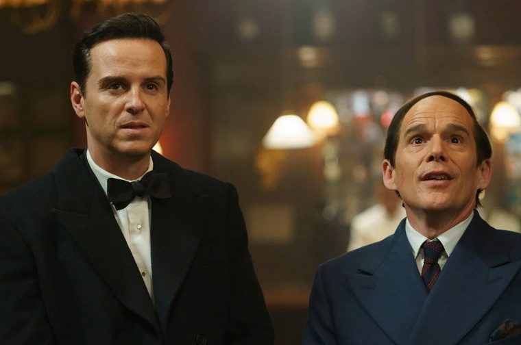 A man in his forties wearing a tuxedo stands next to a balding smaller man in his fifties, who is wearing a blue suit. The setting is period 1940s and we can see a bar and a blurry bartender in the background.