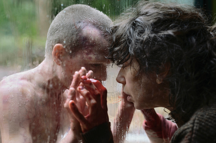 Close up two people, a woman and a child, either side of a rainy window. Their hands and heads are touching through the glass.