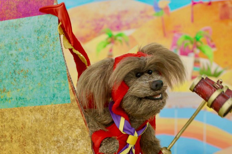 A dog puppet sticks his head out of a multi coloured tent, holding a pair of red and gold binoculars on a stick.