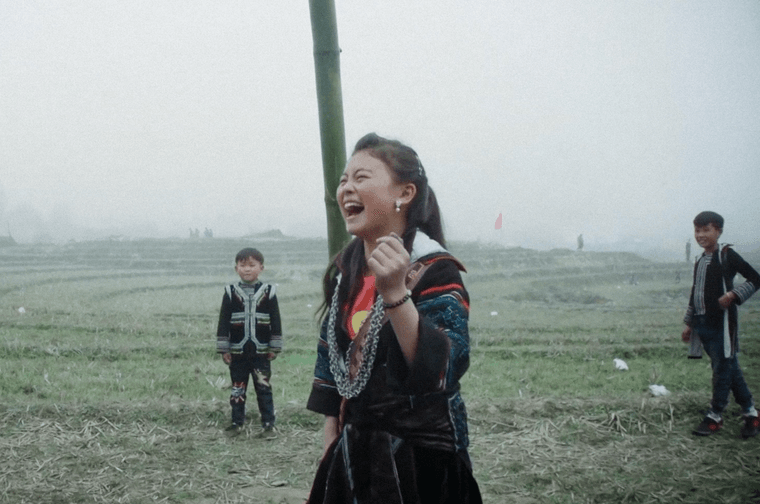Photograph of Vietnamese children playing outside. The young girl at the front has long dark hair and is wearing traditional clothing. She is laughing. Behind her are two young boys, also wearing traditional clothing.