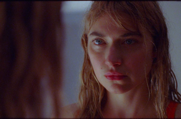 A striking cinematic close-up of a younger woman with wet blonde hair and piercing blue eyes staring at herself in the mirror. The image features dramatic lighting with warm, golden tones illuminating the subject's face against a darker background. The composition creates an intimate, contemplative mood with the direct gaze toward the camera.