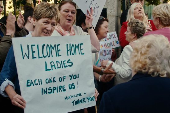 A crowd of older women stand holding placards, one of them reading 'Welcome Home Ladies - Each One You Inspire Us. Some of them are also applauding and cheering.
