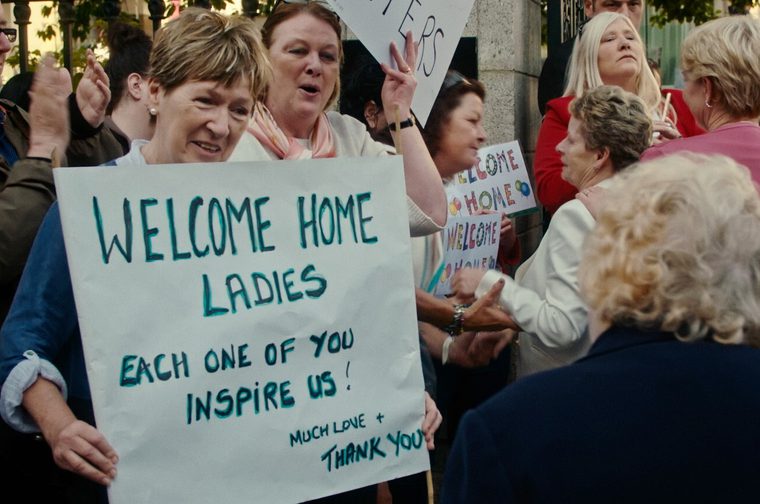 A crowd of older women stand holding placards, one of them reading 'Welcome Home Ladies - Each One You Inspire Us. Some of them are also applauding and cheering.