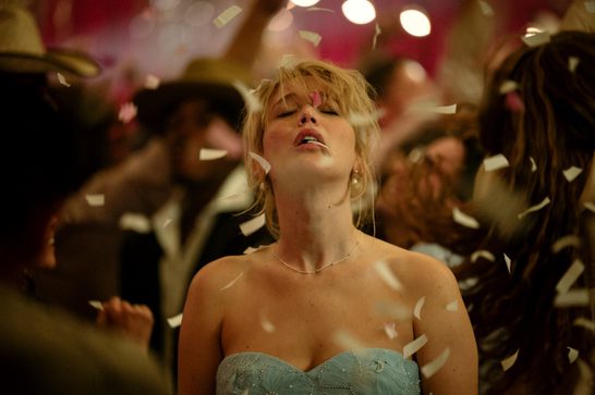 A woman in her thirties with blonde hair (Jennifer Lawrence) stands in the middle of a dance floor, her head raised to the ceiling and her eyes closed. She is wearing a light blue dress and coloured confetti falls around her. We can see blurry images of other people on the dancefloor behind her.