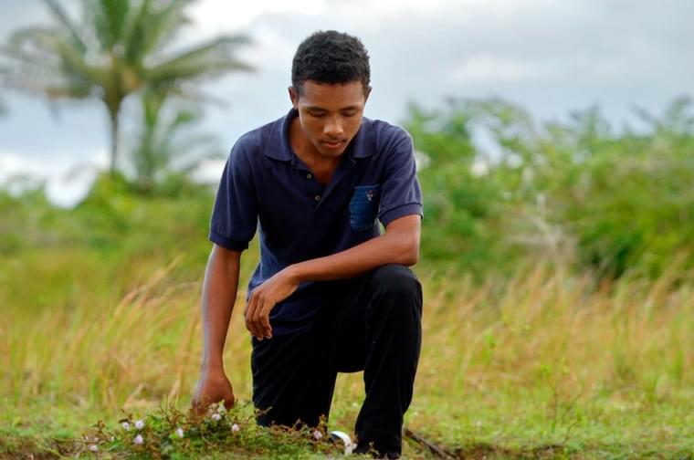 A young Black man wearing a dark blue shirt and black trousers kneels down in some grass, his left arm resting on his left knee. He looks down at the ground in solemn prayer.