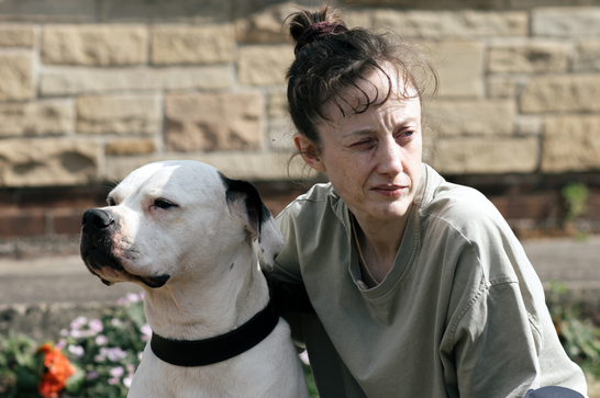 A woman in her late thirties with messy brunette hair tied up in a bob crouches next to a dog, looking off to the right of the image. Behind is a brick wall and a small suburban garden.