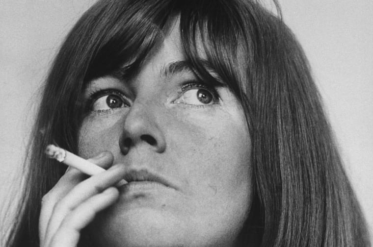 A white women in her twenties with long brunette hair stares off to the right of camera, a lit cigarette held to her lips.