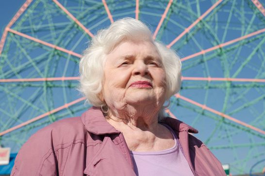We see an elderly woman in her 90s with white, wavy hair, wearing a mauve/dusty pink jacket over a lighter pink top. She's photographed from a low angle looking upward, which gives the image an uplifting, inspirational quality. Behind her is what appears to be a colorful Ferris wheel or similar carnival ride with geometric patterns in coral/pink, green, and blue against a clear blue sky.
