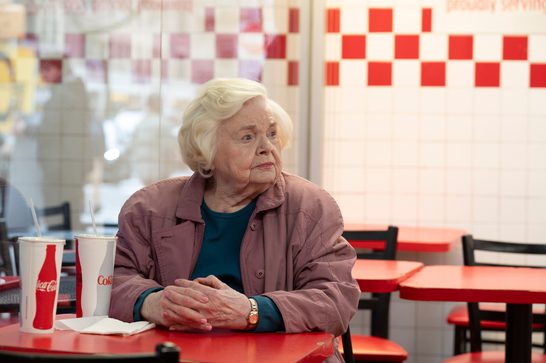 An elderly woman in her 90s sits alone at a red table in what appears to be a fast-food restaurant. She has white hair and is wearing a mauve-colored jacket over a dark teal top. Her hands are clasped together on the table in front of her, and there are drink cups nearby. The setting has a distinctive red and white checkered pattern visible in the background, creating a classic diner or fast-food aesthetic. The woman's expression appears contemplative or perhaps a bit lonely, and the composition emphasizes her solitude in the space.