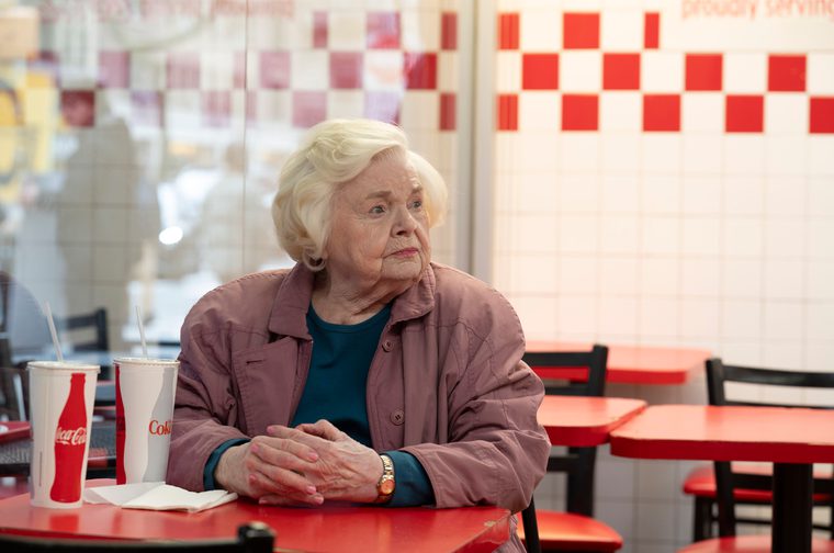 An elderly woman in her 90s sits alone at a red table in what appears to be a fast-food restaurant. She has white hair and is wearing a mauve-colored jacket over a dark teal top. Her hands are clasped together on the table in front of her, and there are drink cups nearby. The setting has a distinctive red and white checkered pattern visible in the background, creating a classic diner or fast-food aesthetic. The woman's expression appears contemplative or perhaps a bit lonely, and the composition emphasizes her solitude in the space.