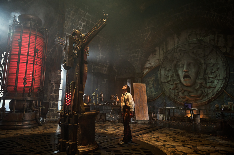 A man in period clothing stands in the centre of a gothic laboratory, looking up at towering brass and copper structure. The atmospheric scene features a large cylindrical apparatus with red lighting on the left, ornate scientific instruments, and stone walls decorated with carved faces and mysterious symbols. Steam or smoke fills the air, and the cobblestone floor and arched ceiling create a dungeon-like atmosphere.