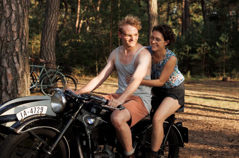 A man in his thirties wearing glasses, a blue vest and orange shorts sits on a motorcycle from the 1940s. A woman of the same age, also wearing glasses, holds onto his waist as she sits behind him. They are both smiling and are driving through a sun dappled forest.
